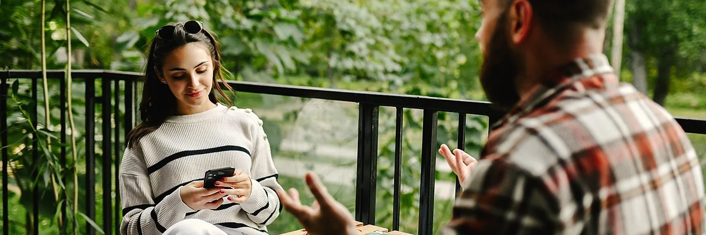 Auf einem Balkon zum Garten sitzen sich eine junge Frau und ein junger Mann an einem Tisch gegenüber. Die Frau liest lächelnd auf ihrem Smartphone. Der Mann hebt gestikulierend beide Hände.