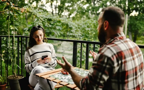 Auf einem Balkon zum Garten sitzen sich eine junge Frau und ein junger Mann an einem Tisch gegenüber. Die Frau liest lächelnd auf ihrem Smartphone. Der Mann hebt gestikulierend beide Hände.
