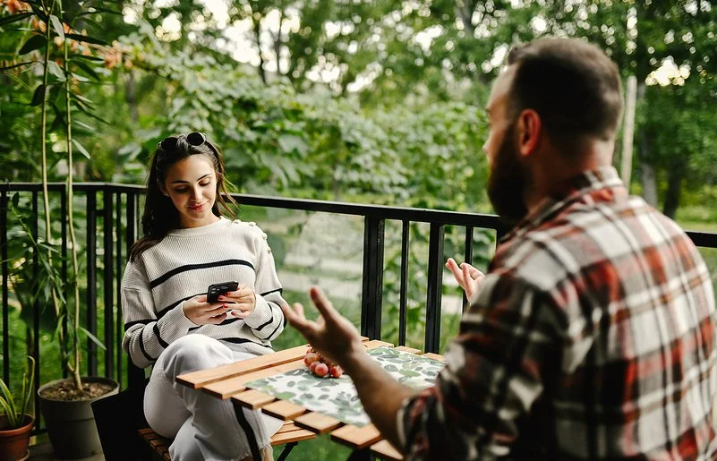 Auf einem Balkon zum Garten sitzen sich eine junge Frau und ein junger Mann an einem Tisch gegenüber. Die Frau liest lächelnd auf ihrem Smartphone. Der Mann hebt gestikulierend beide Hände.
