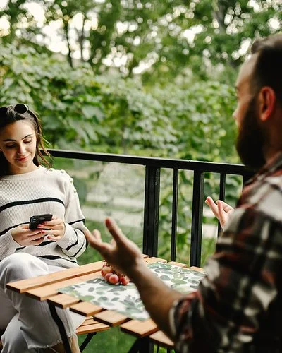 Auf einem Balkon zum Garten sitzen sich eine junge Frau und ein junger Mann an einem Tisch gegenüber. Die Frau liest lächelnd auf ihrem Smartphone. Der Mann hebt gestikulierend beide Hände.