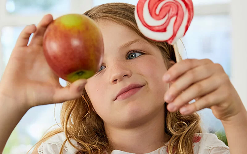 Ein Mädchen hält in einer Hand einen Apfel, in der anderen einen großen runden Lolli und schaut skeptisch auf den Apfel.