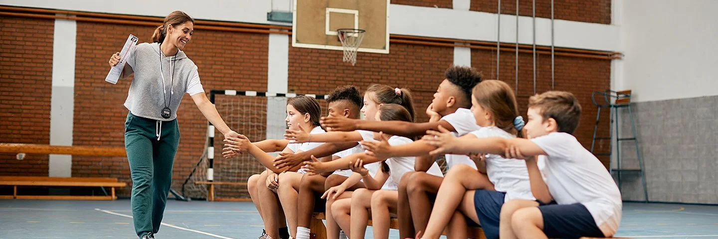 Eine Trainerin oder Sportlehrerin begrüßt eine Gruppe Kinder in Sportkleidung mit Handschlag.