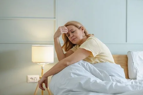 Eine müde aussehende Frau sitzt mit geschlossenen Augen im Bett und fasst sich mit einer Hand an die Stirn.