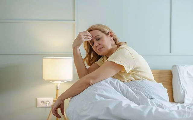 Eine müde aussehende Frau sitzt mit geschlossenen Augen im Bett und fasst sich mit einer Hand an die Stirn.
