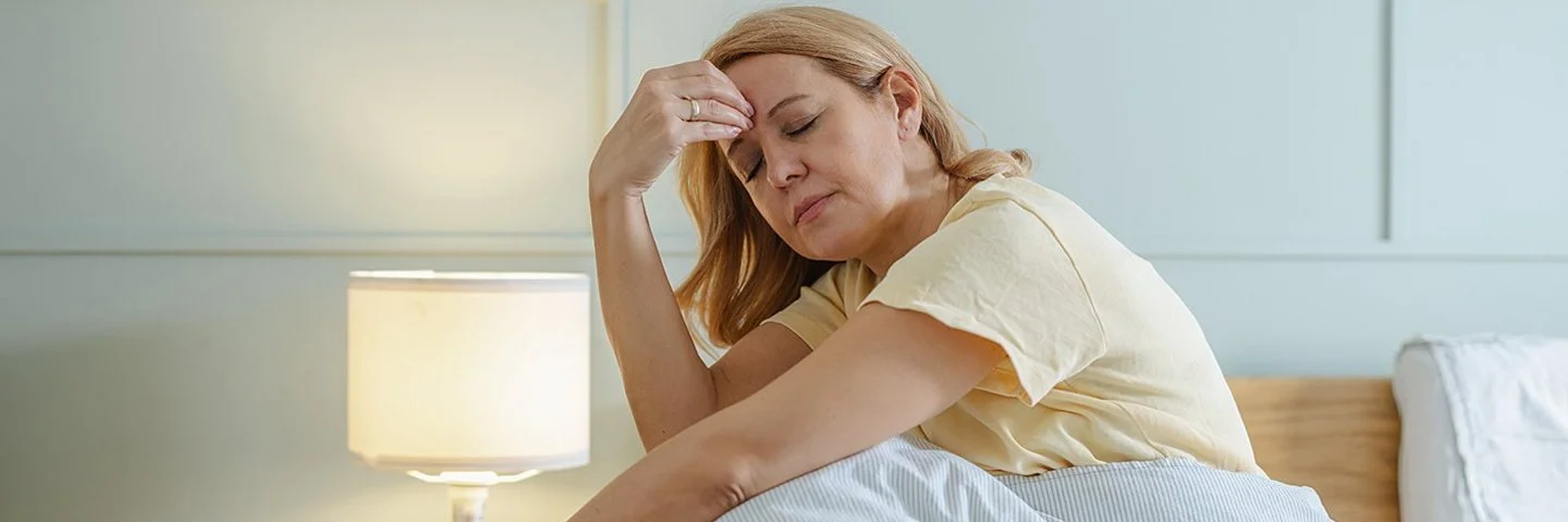 Eine müde aussehende Frau sitzt mit geschlossenen Augen im Bett und fasst sich mit einer Hand an die Stirn.