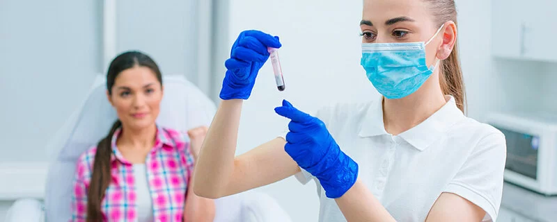 Eine Krankenschwester mit Gesichtsmaske betrachtet eine Blutprobe in einem Reagenzglas, während ihre Patientin sie anschaut.