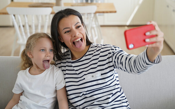 Mutter und Tochter sitzen im Wohnzimmer auf der Couch und machen ein Selfie, während sie ihre Zungen rausstrecken.