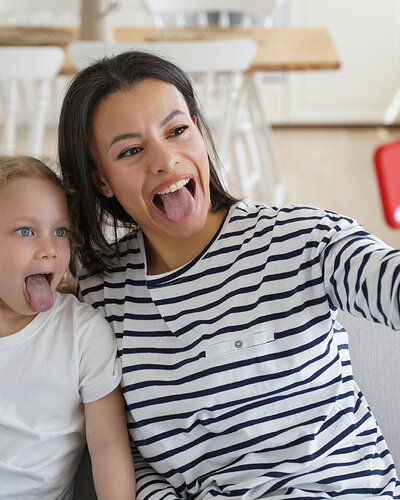 Mutter und Tochter sitzen im Wohnzimmer auf der Couch und machen ein Selfie, während sie ihre Zungen rausstrecken.