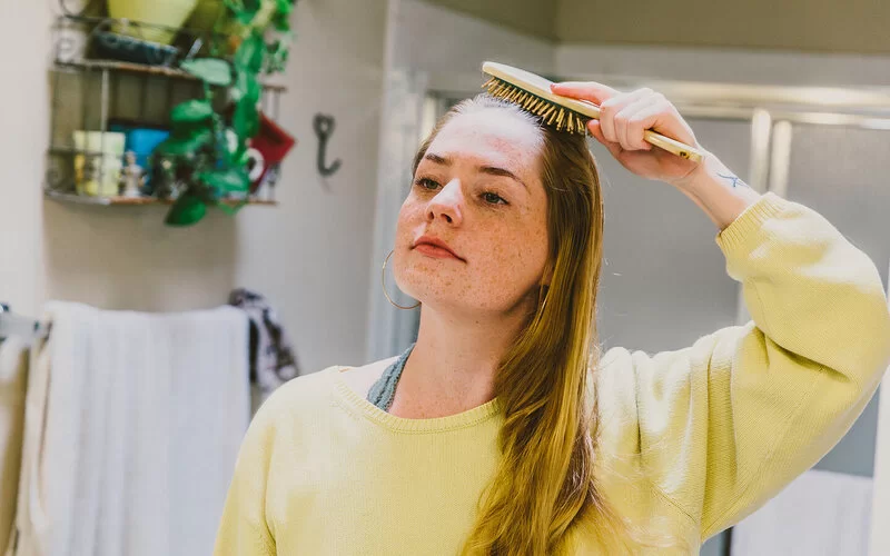 Eine junge Frau steht in einem Badezimmer. In der rechten Hand hält sie eine Haarbürste, mit der sie sich durch die Haare fährt.