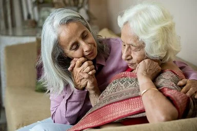 Eine Frau hält die Hand ihrer alten Mutter. Beide sitzen in einem Wohnzimmer auf einer beigefarbenen Couch. Im Hintergrund ist ein Bücherregal zu sehen.