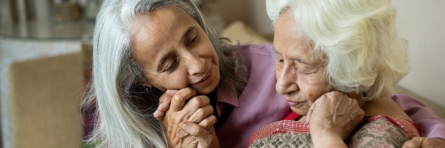 Eine Frau hält die Hand ihrer alten Mutter. Beide sitzen in einem Wohnzimmer auf einer beigefarbenen Couch. Im Hintergrund ist ein Bücherregal zu sehen.