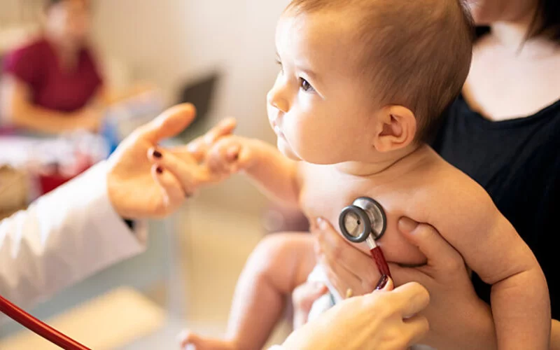 Eine Mutter hält ihr Baby mit Pseudokrupp auf dem Arm, während eine Ärztin ihm die Lunge abhört.