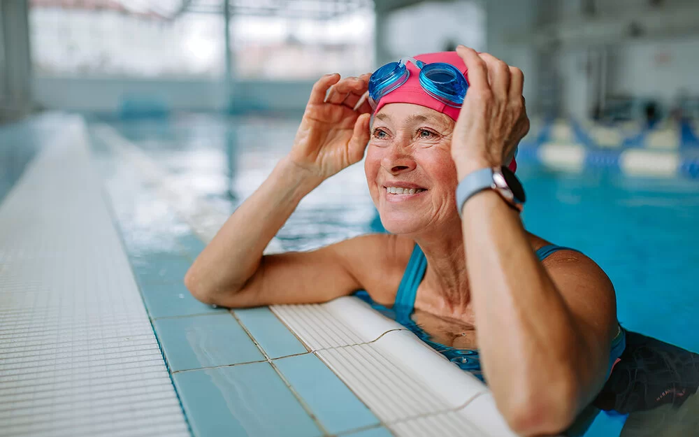 Eine Frau im Alter von etwa 60 Jahren stützt sich lächelnd am Beckenrand eines Schwimmbads auf und berührt seitlich ihre Schwimmbrille, die sie nach oben über ihre Badekappe geschoben hat.