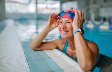 Eine Frau im Alter von etwa 60 Jahren stützt sich lächelnd am Beckenrand eines Schwimmbads auf und berührt seitlich ihre Schwimmbrille, die sie nach oben über ihre Badekappe geschoben hat.