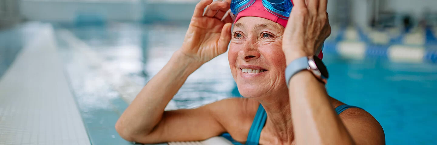 Eine Frau im Alter von etwa 60 Jahren stützt sich lächelnd am Beckenrand eines Schwimmbads auf und berührt seitlich ihre Schwimmbrille, die sie nach oben über ihre Badekappe geschoben hat.