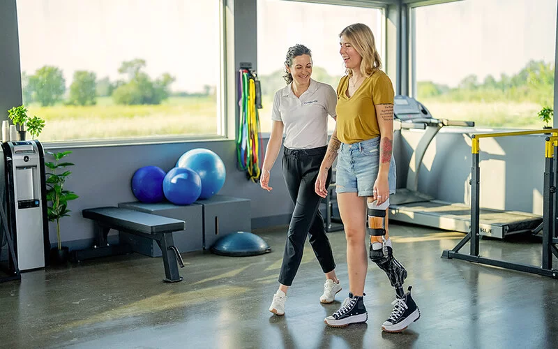 In einer physiotherapeutischen Praxis führt eine junge Frau mit einem amputierten linken Bein und einer Beinprothese Laufübungen durch. Eine Physiotherapeutin geht neben ihr und unterstützt sie dabei.