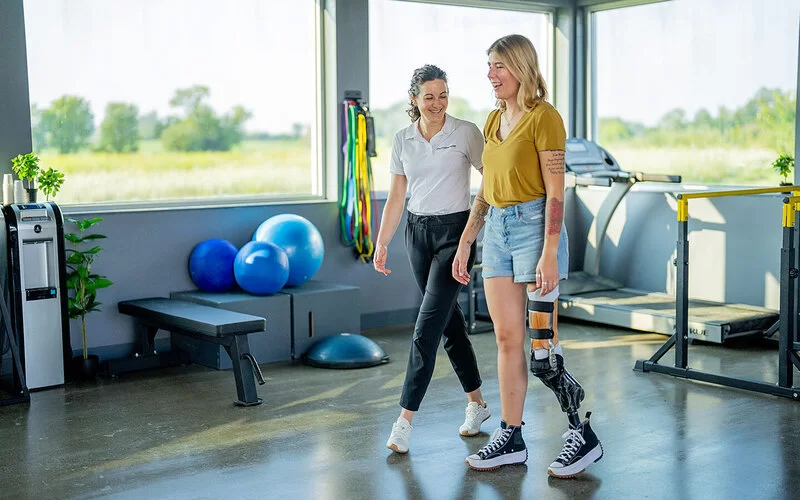 In einer physiotherapeutischen Praxis führt eine junge Frau mit einem amputierten linken Bein und einer Beinprothese Laufübungen durch. Eine Physiotherapeutin geht neben ihr und unterstützt sie dabei.