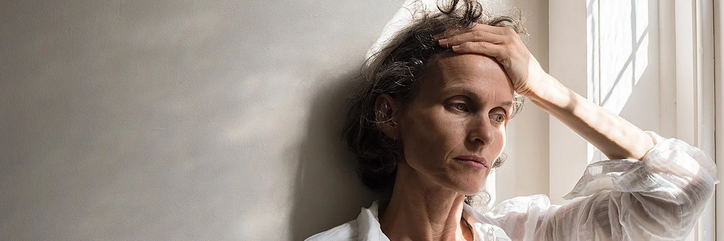 Eine ältere nachdenklich und angespannt wirkende Frau lehnt mit dem Rücken an einer Wand neben einem Fenster, während sie sich mit der linken Hand an den Kopf fasst.