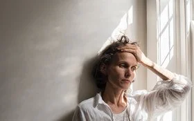 Eine ältere nachdenklich und angespannt wirkende Frau lehnt mit dem Rücken an einer Wand neben einem Fenster, während sie sich mit der linken Hand an den Kopf fasst.