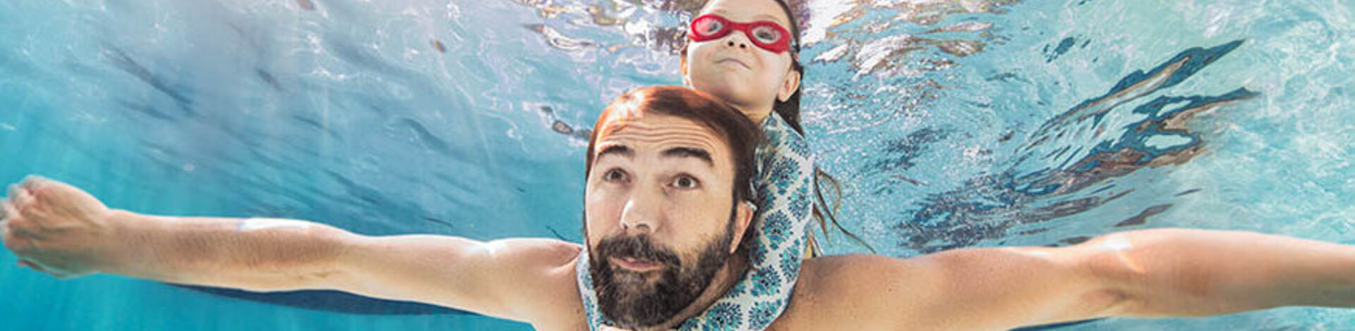 Ein Mann schwimmt unter Wasser. Ein Kind hält sich an seinen Schultern fest. 
