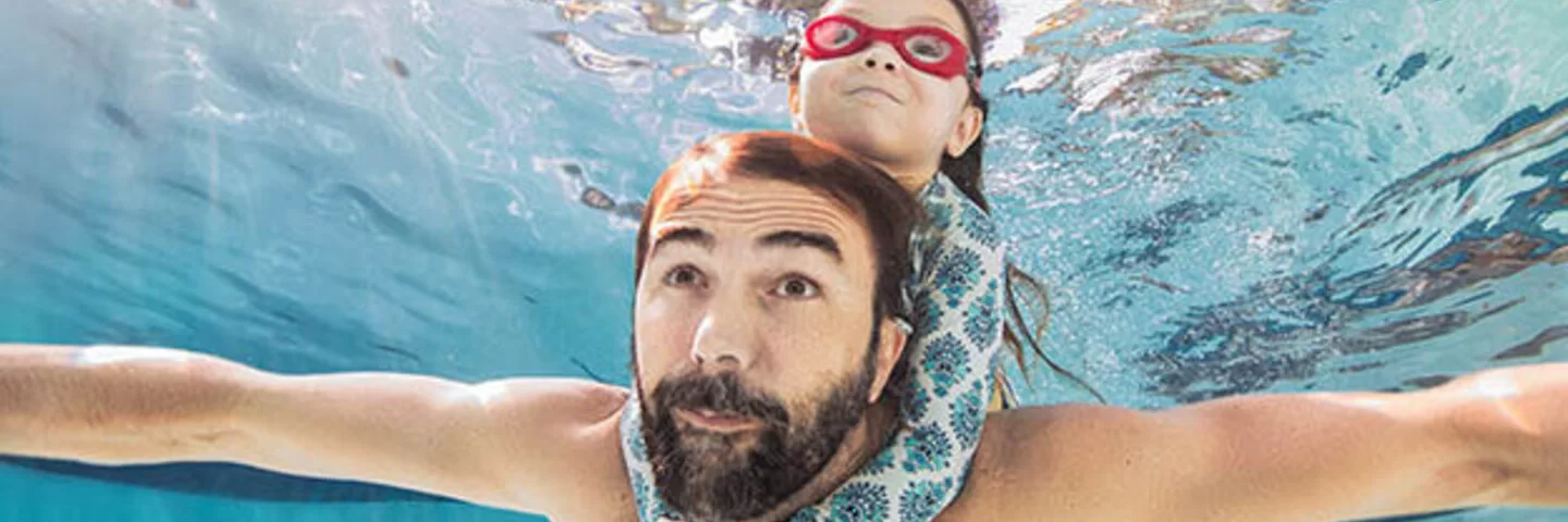 Ein Mann schwimmt unter Wasser. Ein Kind hält sich an seinen Schultern fest. 
