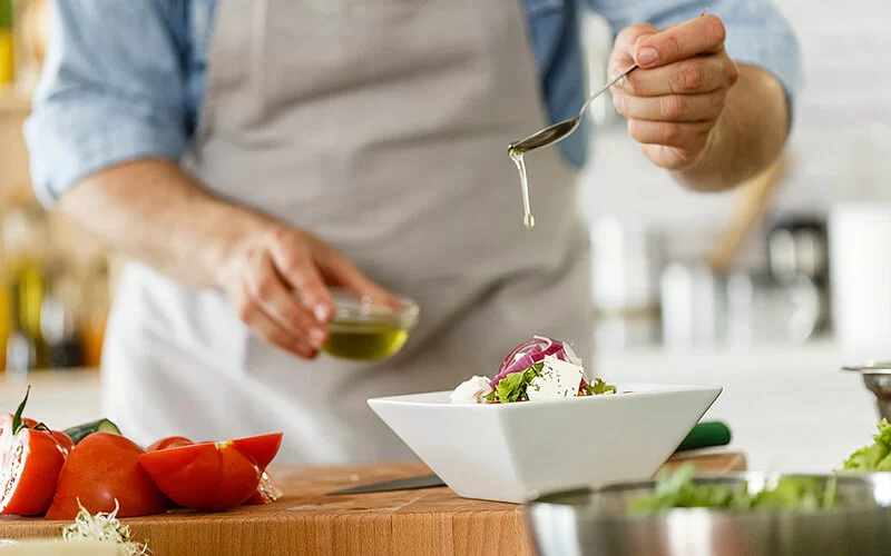Eine nicht erkennbare Person gibt mit einem Esslöffel Öl auf einen Salat in einer viereckigen weißen Schüssel.