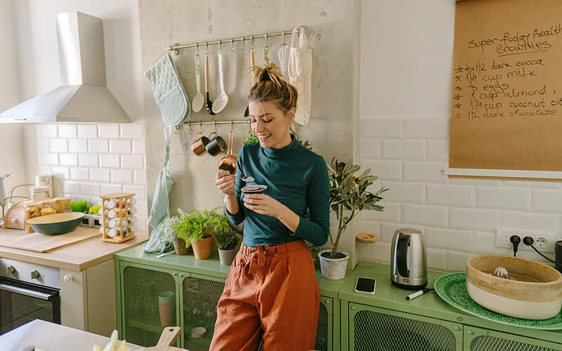 Eine junge Frau lehnt an einem Sideboard in ihrer Küche und löffelt entspannt einen Joghurt.