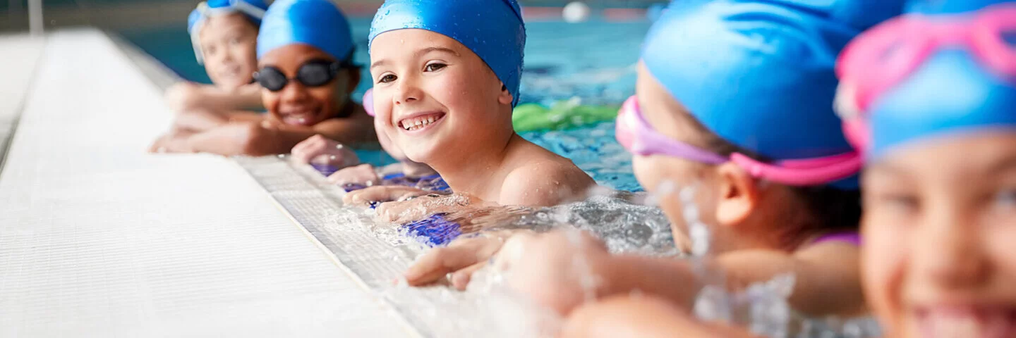 Kinder mit Badekappen und Schwimmbrillen halten sich am Beckenrand fest.