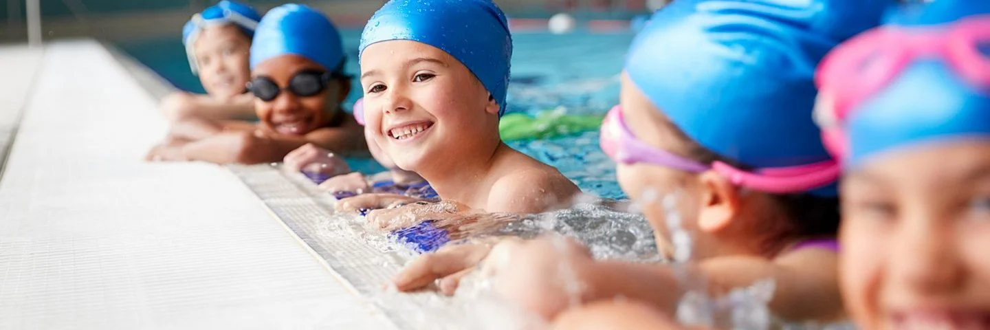 Kinder mit Badekappen und Schwimmbrillen halten sich am Beckenrand fest.