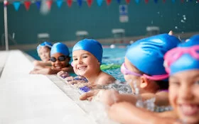 Kinder mit Badekappen und Schwimmbrillen halten sich am Beckenrand fest.