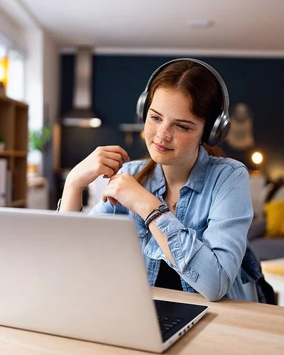 Ein Mädchen im Teenageralter sitzt mit Kopfhörern vor einem Laptop an einem Tisch im Wohnzimmer. Lächelnd blickt sie auf den Bildschirm.