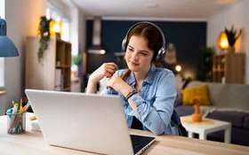 Ein Mädchen im Teenageralter sitzt mit Kopfhörern vor einem Laptop an einem Tisch im Wohnzimmer. Lächelnd blickt sie auf den Bildschirm.