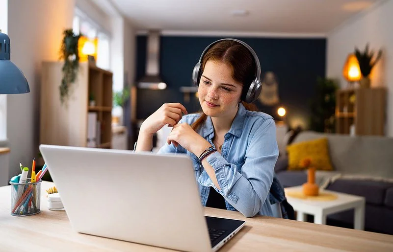 Ein Mädchen im Teenageralter sitzt mit Kopfhörern vor einem Laptop an einem Tisch im Wohnzimmer. Lächelnd blickt sie auf den Bildschirm.