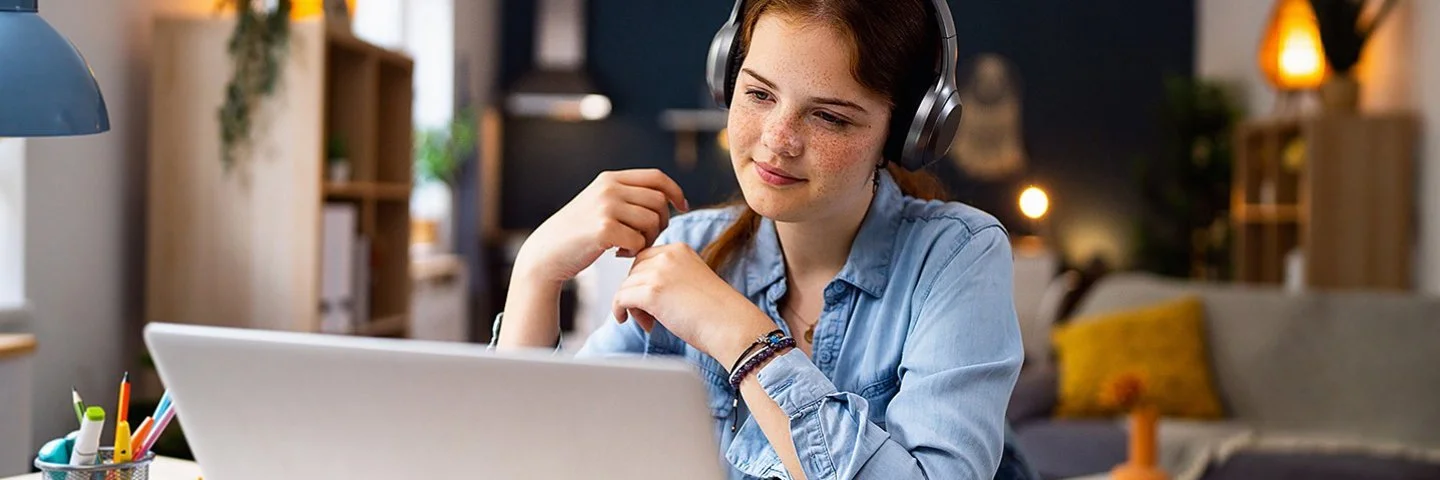 Ein Mädchen im Teenageralter sitzt mit Kopfhörern vor einem Laptop an einem Tisch im Wohnzimmer. Lächelnd blickt sie auf den Bildschirm.