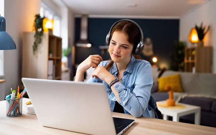 Ein Mädchen im Teenageralter sitzt mit Kopfhörern vor einem Laptop an einem Tisch im Wohnzimmer. Lächelnd blickt sie auf den Bildschirm.