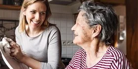 Eine junge und eine ältere Frau stehen gemeinsam in der Küche an einer Spüle. Sie unterhalten sich lachend, während die jüngere Frau das Geschirr abtrocknet. Im Hintergrund sind verschiedene Küchenmöbel, Regale und Küchenutensilien zu sehen.