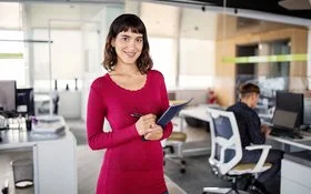 Eine junge Frau mit schulterlangem Haar steht in einem Büro und blickt selbstbewusst in die Kamera. Im Hintergrund sind mehrere Schreibtische zu sehen, an einem der Schreibtische sitzt ein Mann mittleren Alters auf einem Bürostuhl.