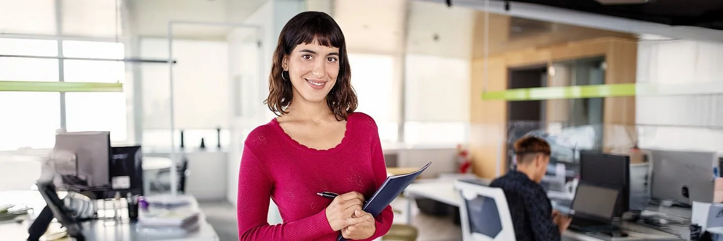 Eine junge Frau mit schulterlangem Haar steht in einem Büro und blickt selbstbewusst in die Kamera. Im Hintergrund sind mehrere Schreibtische zu sehen, an einem der Schreibtische sitzt ein Mann mittleren Alters auf einem Bürostuhl.