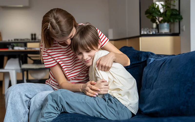 Eine Mutter umarmt ihren Sohn von hinten auf dem Sofa, beide lachen herzlich.