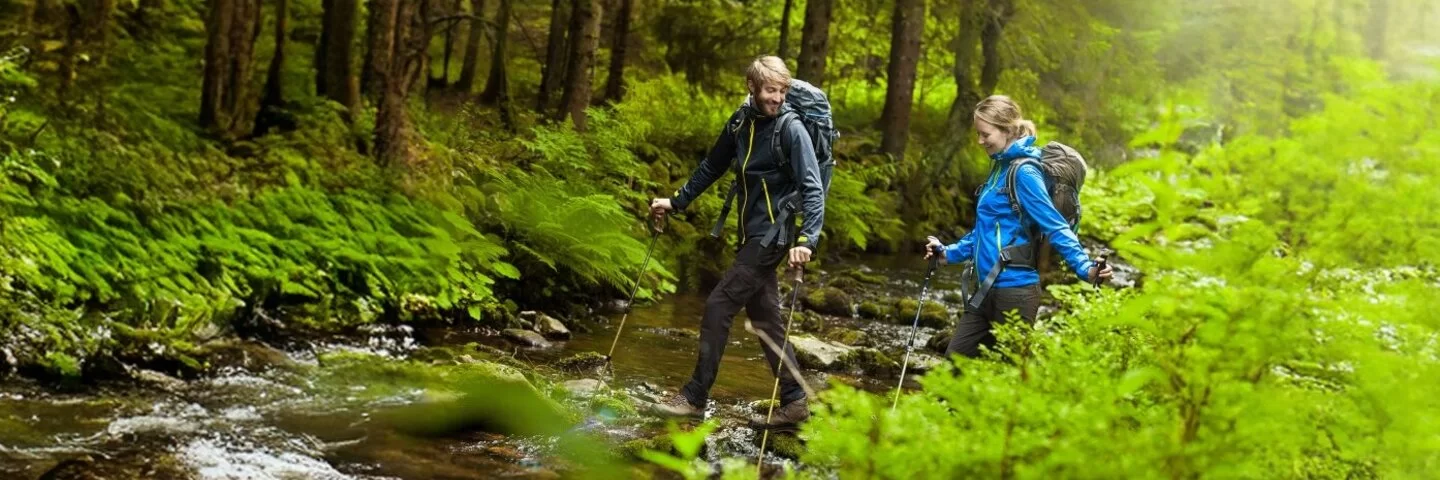 Junges Paar wandert mit Nordic Walking Stöcken im idyllischen Wald durch einen Bach.