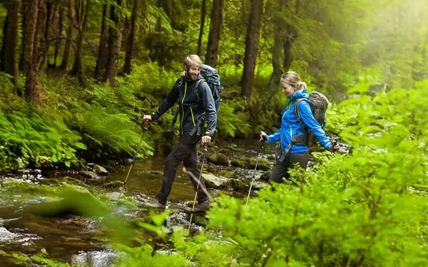 Junges Paar wandert mit Nordic Walking Stöcken im idyllischen Wald durch einen Bach.