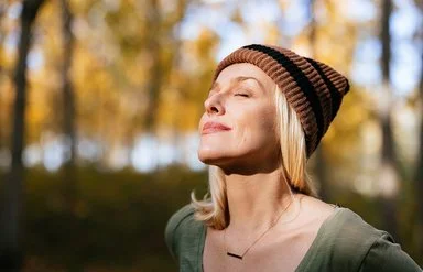 Porträtfoto einer jüngeren Frau mit blonden Haaren und einer Mütze, die im Wald steht und das Sonnenlicht genießt.