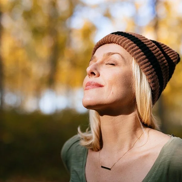 Porträtfoto einer jüngeren Frau mit blonden Haaren und einer Mütze, die im Wald steht und das Sonnenlicht genießt.