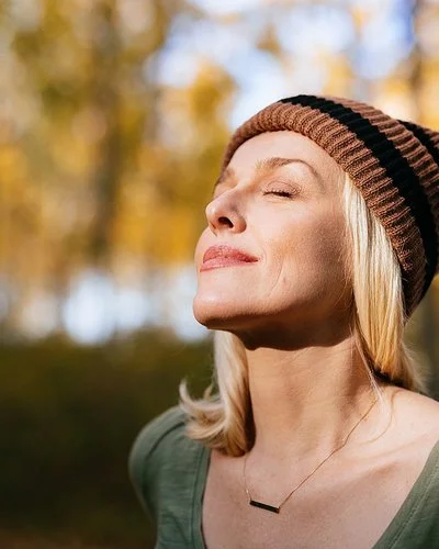 Porträtfoto einer jüngeren Frau mit blonden Haaren und einer Mütze, die im Wald steht und das Sonnenlicht genießt.