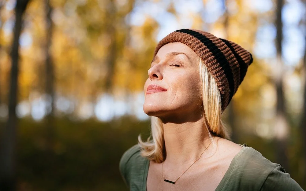 Porträtfoto einer jüngeren Frau mit blonden Haaren und einer Mütze, die im Wald steht und das Sonnenlicht genießt.