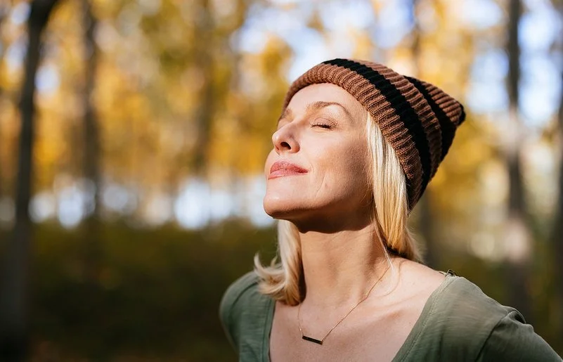 Porträtfoto einer jüngeren Frau mit blonden Haaren und einer Mütze, die im Wald steht und das Sonnenlicht genießt.