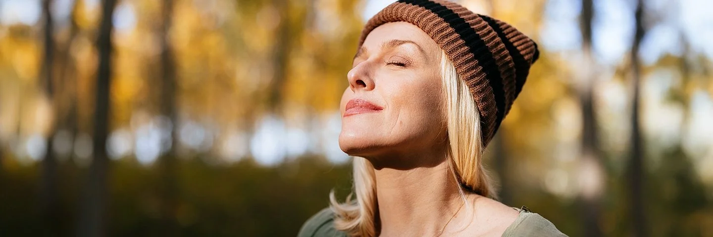 Porträtfoto einer jüngeren Frau mit blonden Haaren und einer Mütze, die im Wald steht und das Sonnenlicht genießt.