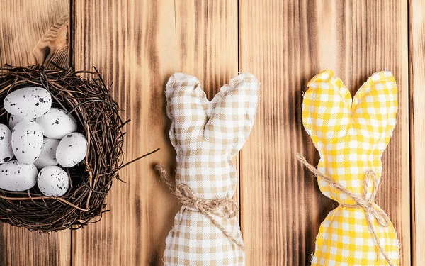 Zwei selbstgenähte Osterhasen aus kariertem Stoff liegen neben einem kleinen Osternest, das mit Wachteleiern befüllt ist.