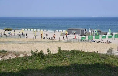 Menschen spielen am Strand Beachvolleyball, während andere sich im Sand entspannen. Im Hintergrund ist das Meer zu sehen.