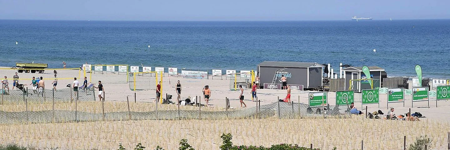 Menschen spielen am Strand Beachvolleyball, während andere sich im Sand entspannen. Im Hintergrund ist das Meer zu sehen.